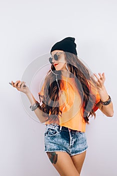 Young hipster beautiful woman in sunglasses wearing in black hat and orange T-shirt listening music in headphones near