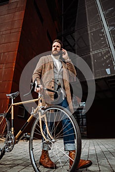 Young hipster businessman walking with his bike and talking on phone. Stylish manager in business area. Outside
