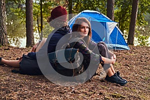 Young hipster couple hiking in the forest and sitting back to back while warming near a campfire at camp.