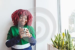 Young hipster dark-skinned woman dressed in casual clothes holding cup of hot drink, enjoying coffee or tea