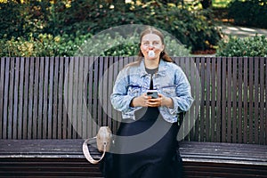 Young hipster female relaxing on the bench in the park.. Model blowing bubble with chewing gum. Happy 30s Woman looking