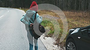 Young hipster girl getting out of car with backpack and hiking right along highway or road. Woman in red hat traveling by
