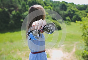 Young hipster girl is travelling in the mountains. Woman in modern dress is holding fashion sunglasses in hands. Trees and sky