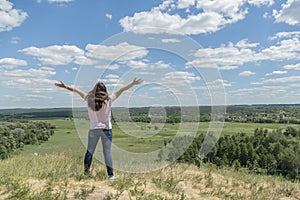 Young hipster girl is travelling in summer holidays. Woman in modern dress is standing on the hill and looking at the field. Blue