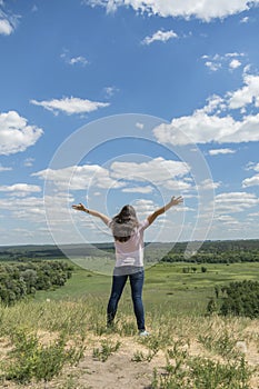 Young hipster girl is travelling in summer holidays. Woman in modern dress is standing on the hill and looking at the field. Blue