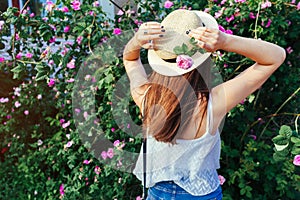 Young hipster girl wearing hat walking by blooming roses. Woman enjoys flowers in park. Summer outfit.