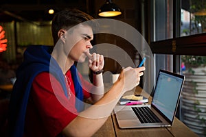 Young hipster guy with thoughtful look checking e-mail on mobile phone while sitting with laptop computer