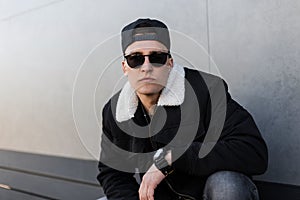 Young hipster man in a black cap in trendy sunglasses in a vintage black jacket with fur in gray ripped jeans