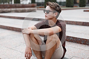 Young hipster man in black sunglasses with a stylish hairstyle in a brown t-shirt in summer shorts sits on the vintage steps