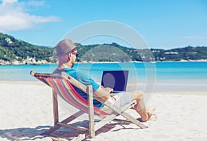 Young hipster man with laptop on tropical beach. Travel, vacation, internet, freelance job concept photo