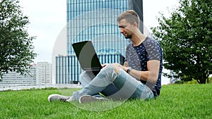 Young hipster man sitting on grass and use laptop, typing on keyboard, modern business glass building on background