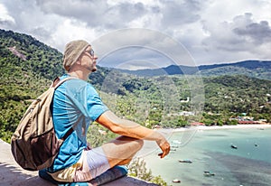Young hipster man sitting at sea view point with backpack. Travel, adventure, wonderlust, tourism concept.