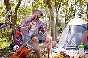 Young hipster man smiling, sitting on chair and enjoying camping in nature. Picnic in forest. Holiday, leisure, fun, lifestyle