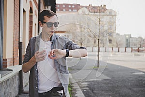 Young hipster man walking on the street, looking at watch and using his smartphone. With copy space