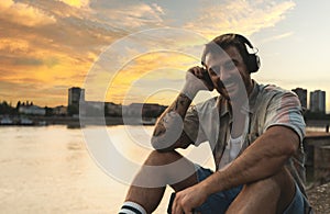 Young hipster millennial man sitting on riverside looking at city listening music on wireless headphones enjoying moment, life and