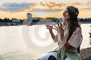 Young hipster millennial woman sitting on riverside looking at city listening music on wireless headphones enjoying moment, life