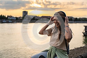 Young hipster millennial woman sitting on riverside looking at city listening music on wireless headphones enjoying moment, life