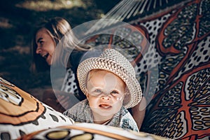 Young hipster mother playing with her cute little son on hammock in summer sunny park. stylish happy mom with kid relaxing in