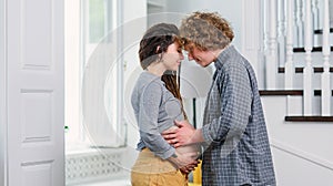 Young hipster pregnant couple standing opposite each other in contemporary apartment and tenderly stroking her belly.