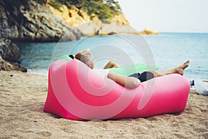 Young hipster relaxing on coastline beach on inflatable lazy air pouffe sofa, person tourist enjoy sunny day on background coast