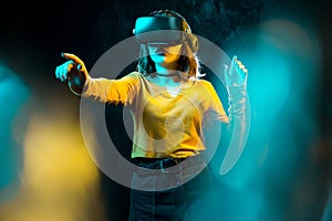 Young hipster woman with curly hair wearing virtual reality goggles and touch another world in studio neon lights