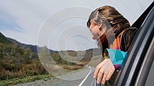 Young hipster woman leaning out of car window and enjoy the vacation. Beautiful girl sitting in car passenger seat