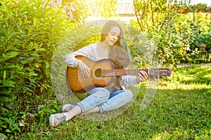 Young hipster woman sitting in grass and playing guitar on park or garden background. Teen girl learning to play song