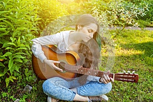 Young hipster woman sitting in grass and playing guitar on park or garden background. Teen girl learning to play song