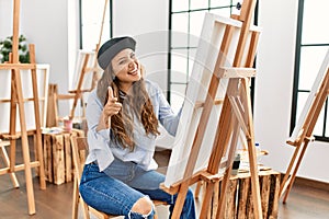 Young hispanic artist woman painting on canvas at art studio pointing fingers to camera with happy and funny face