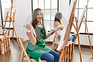 Young hispanic artist women painting on canvas at art studio with a big smile on face, pointing with hand finger to the side