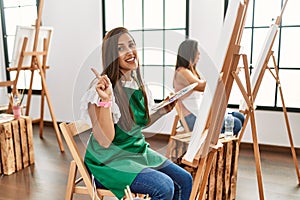 Young hispanic artist women painting on canvas at art studio cheerful with a smile on face pointing with hand and finger up to the
