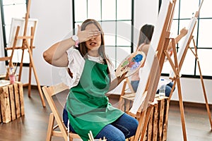 Young hispanic artist women painting on canvas at art studio covering eyes with hand, looking serious and sad