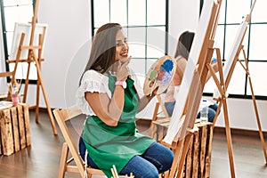 Young hispanic artist women painting on canvas at art studio with hand on chin thinking about question, pensive expression