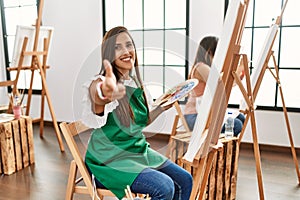 Young hispanic artist women painting on canvas at art studio looking confident at the camera smiling with crossed arms and hand