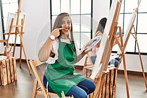 Young hispanic artist women painting on canvas at art studio pointing with hand finger to face and nose, smiling cheerful