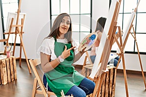 Young hispanic artist women painting on canvas at art studio pointing with hand finger to the side showing advertisement, serious