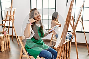 Young hispanic artist women painting on canvas at art studio pointing to the back behind with hand and thumbs up, smiling