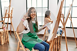 Young hispanic artist women painting on canvas at art studio smiling doing phone gesture with hand and fingers like talking on the