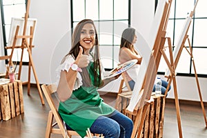 Young hispanic artist women painting on canvas at art studio smiling positive doing ok sign with hand and fingers