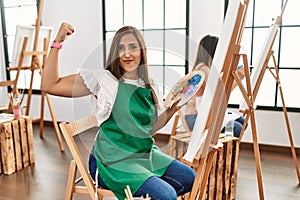 Young hispanic artist women painting on canvas at art studio strong person showing arm muscle, confident and proud of power