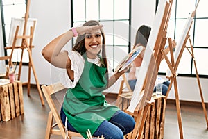Young hispanic artist women painting on canvas at art studio very happy and smiling looking far away with hand over head