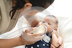 Young hispanic baby or asian infant boy drinking milk from plastic bottle feeding from young parents mother or babysitter