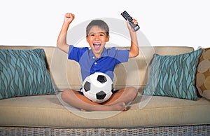 Young hispanic boy happy and excited watching football game on television at home living room couch celebrating scoring goal gestu