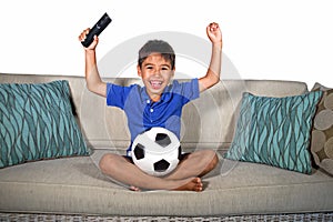 Young hispanic boy happy and excited watching football game on television at home living room couch celebrating scoring goal gestu