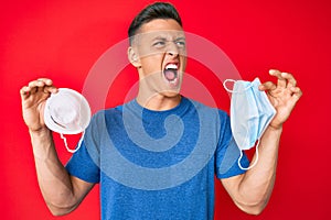 Young hispanic boy holding two diferent coronavirus protection mask angry and mad screaming frustrated and furious, shouting with