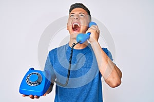 Young hispanic boy holding vintage telephone angry and mad screaming frustrated and furious, shouting with anger looking up