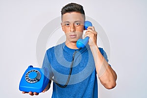 Young hispanic boy holding vintage telephone relaxed with serious expression on face
