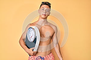 Young hispanic boy shirtless wearing swimwear and holding weighing machine thinking attitude and sober expression looking self