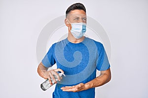 Young hispanic boy wearing medical mask holding hand sanitizer gel angry and mad screaming frustrated and furious, shouting with