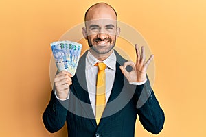 Young hispanic business man holding south african 100 rand banknotes doing ok sign with fingers, smiling friendly gesturing